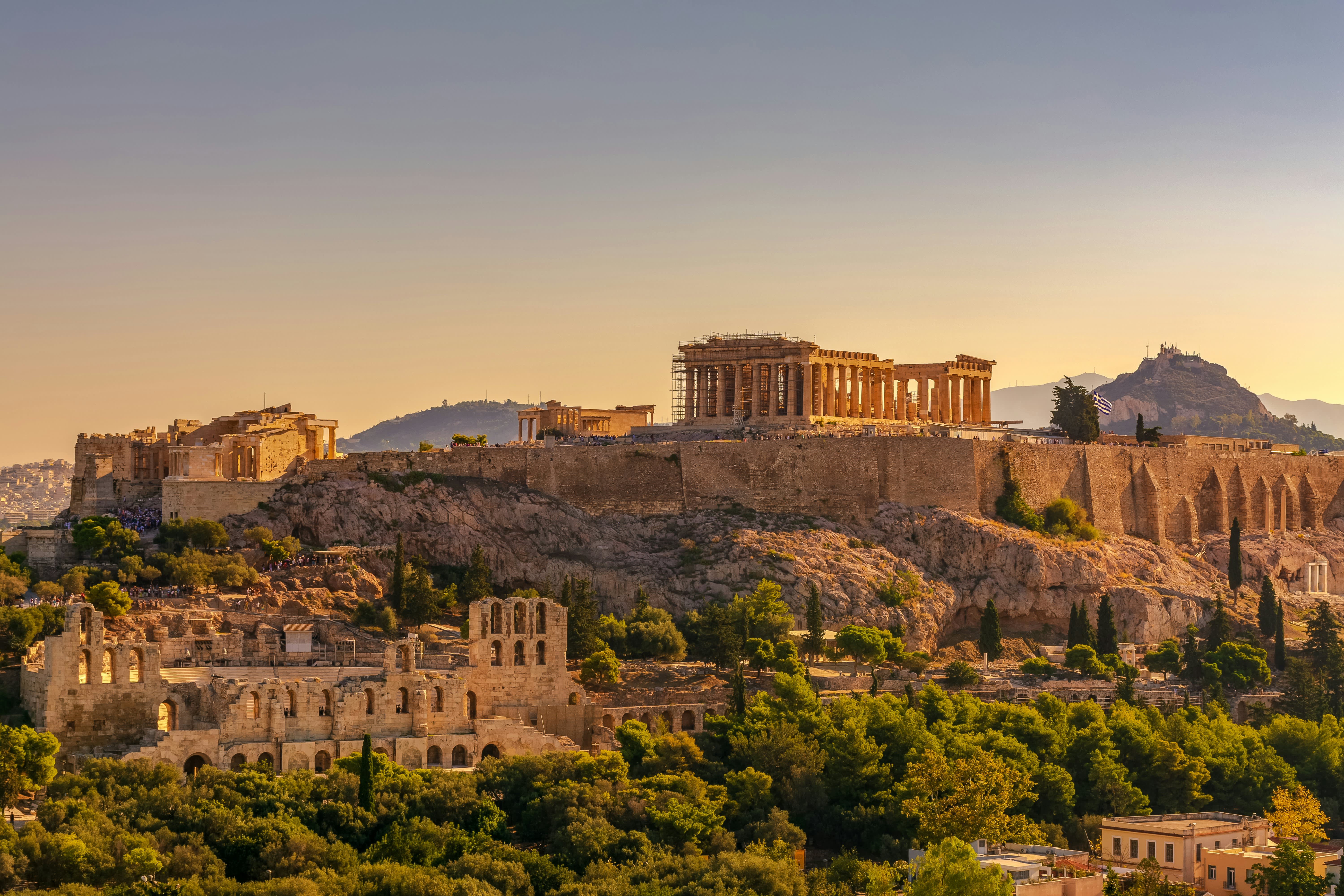 Ancient ruins and lush greenery fill the foreground, while the majestic Acropolis stands prominently on a rocky hill against a clear, pale sky in the background.