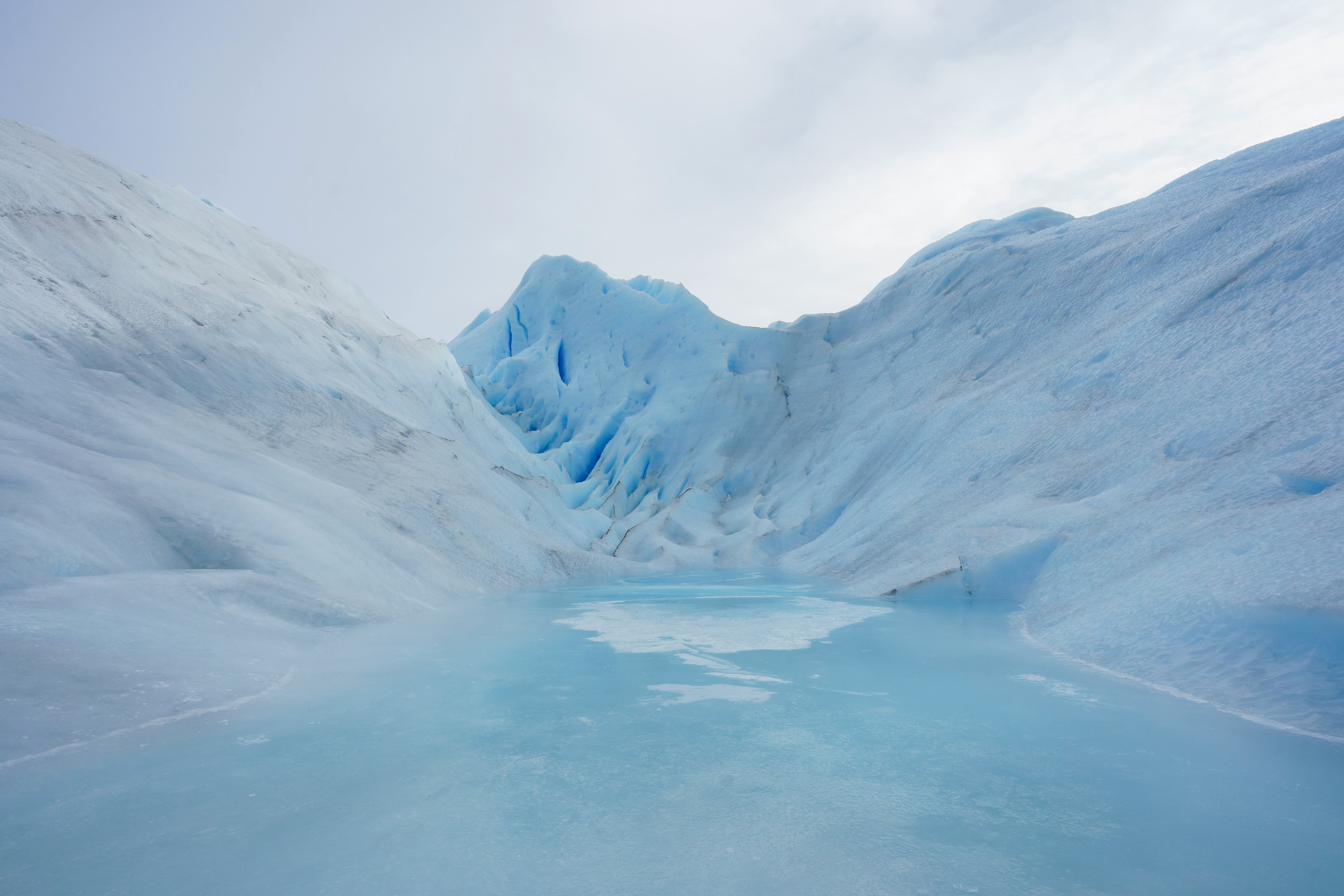 A calm, icy blue lake reflects the towering, sculpted ice formations of a glacier under a pale sky.