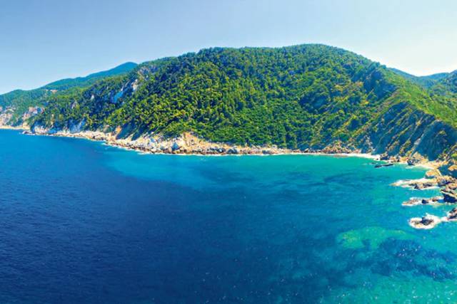 Deep blue ocean meets a turquoise bay with rocky shores, leading to a lush green, tree-covered mountain range under a clear blue sky.