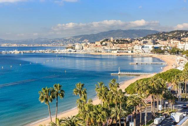 Palm trees line a busy street and beach in the foreground, leading to a sparkling blue bay and a distant city nestled against rolling hills under a bright blue sky.