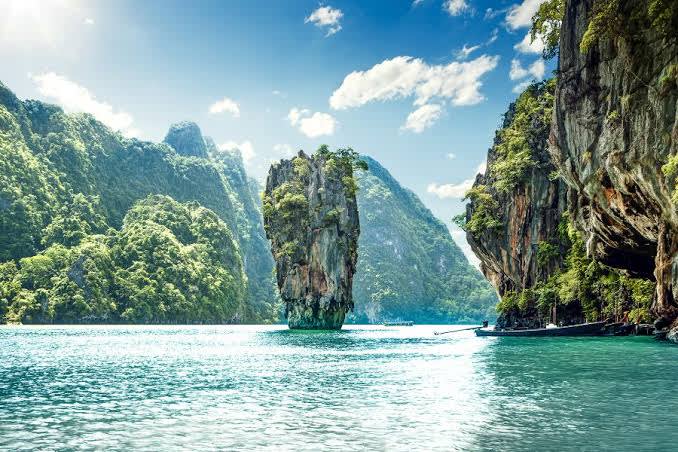 A solitary, tree-topped rock formation rises from the turquoise water in the foreground, while lush, green mountains form the dramatic backdrop under a bright, cloudy sky.
