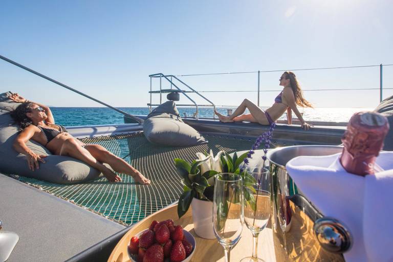 Two women lounge on a catamaran's net trampoline, enjoying a sunny day at sea with champagne and strawberries.