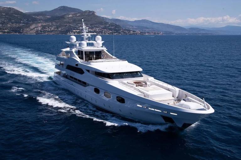 A luxurious white superyacht glides through the deep blue ocean with a mountainous coastline in the background.