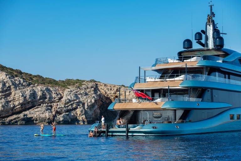 A luxurious turquoise yacht anchors near a rocky coastline as people enjoy paddleboarding and relaxing on deck.