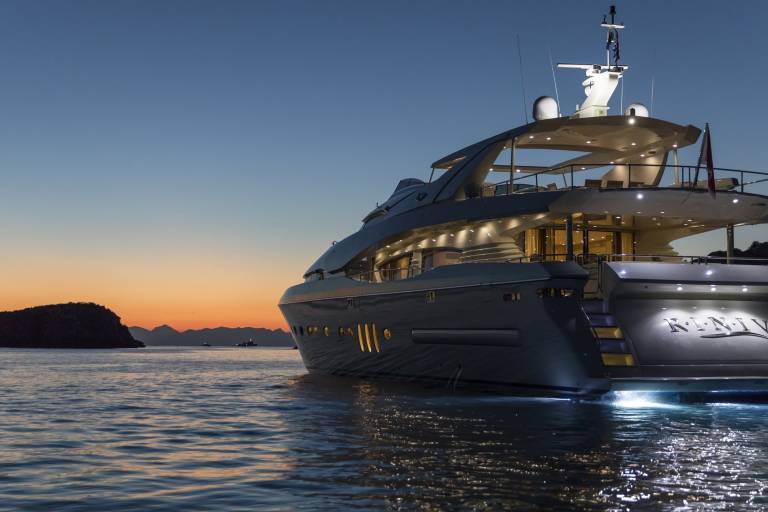 A luxurious yacht named "Rin-IV" is illuminated at dusk as it rests on calm waters near a silhouetted island.