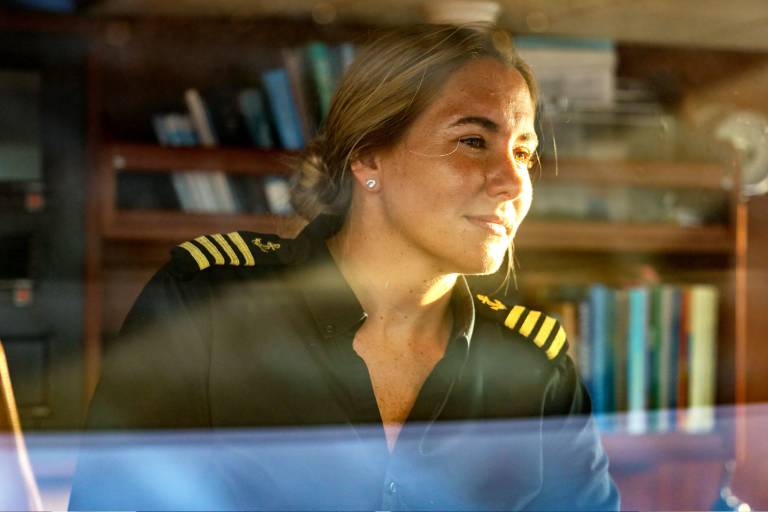 A woman in a pilot's uniform stands in front of a bookshelf, her expression thoughtful and focused.