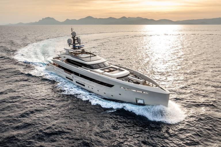 A sleek, modern yacht glides through the shimmering sea at sunset, leaving a trail of white wake against a backdrop of distant mountains.