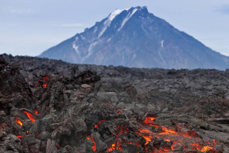 Fiery lava flows cascade down the slopes of a snow-capped volcano under a pale sky.