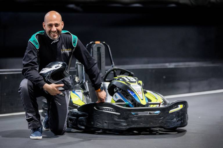 A smiling man in a racing suit kneels beside a go-kart, ready for the track.