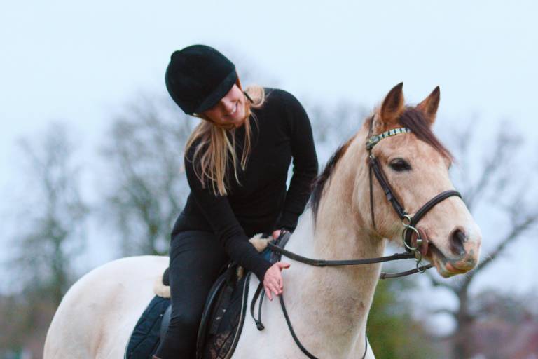 A smiling rider in a black helmet and outfit gently pats her light-colored horse as they stand in a field.