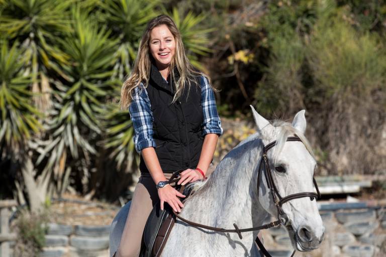 A smiling woman in a plaid shirt and vest rides a white horse through a sunny outdoor arena.