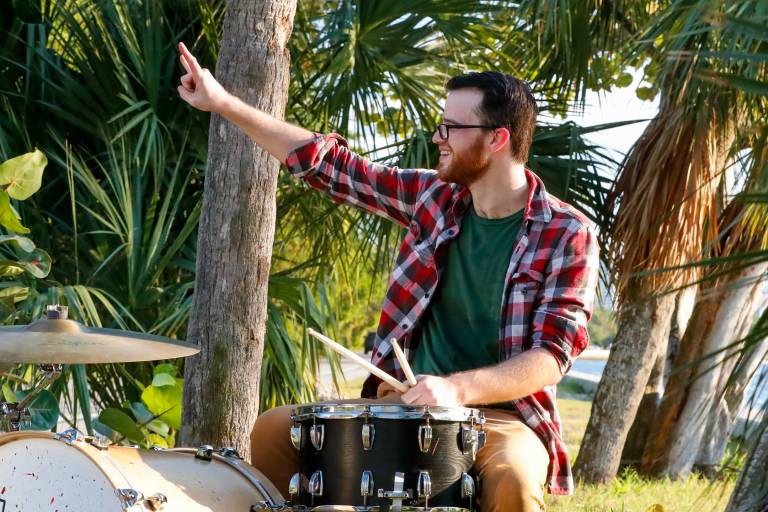 A man with glasses and a plaid shirt plays drums outdoors surrounded by palm trees.
