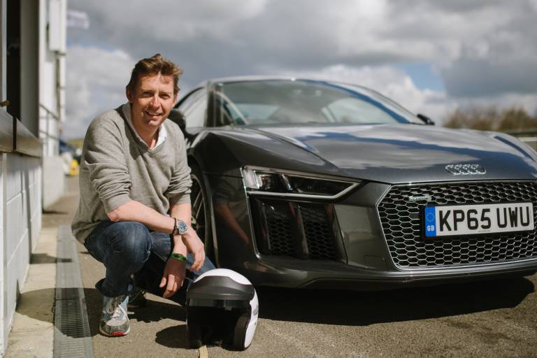 A man in a grey sweater and jeans crouches beside a sleek grey Audi R8 on a sunny day.