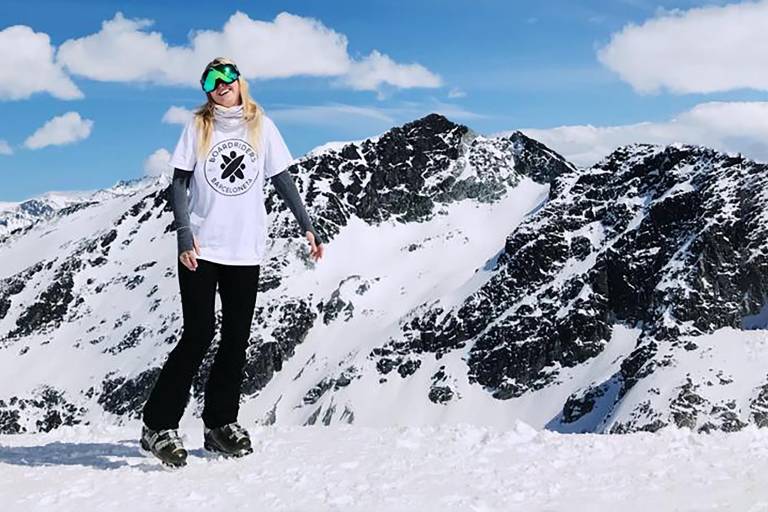 A snowboarder smiles brightly in ski goggles against a backdrop of snow-covered mountains under a blue sky.