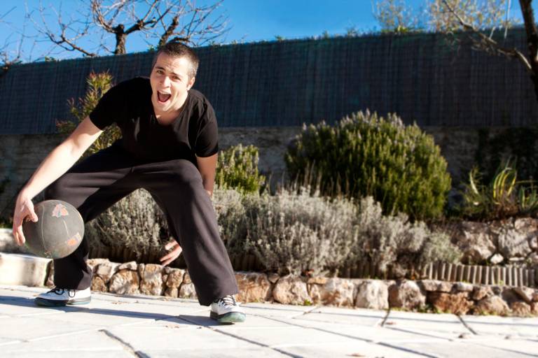 A man in black athletic wear enthusiastically dribbles a basketball on a sun-drenched patio surrounded by lush greenery.