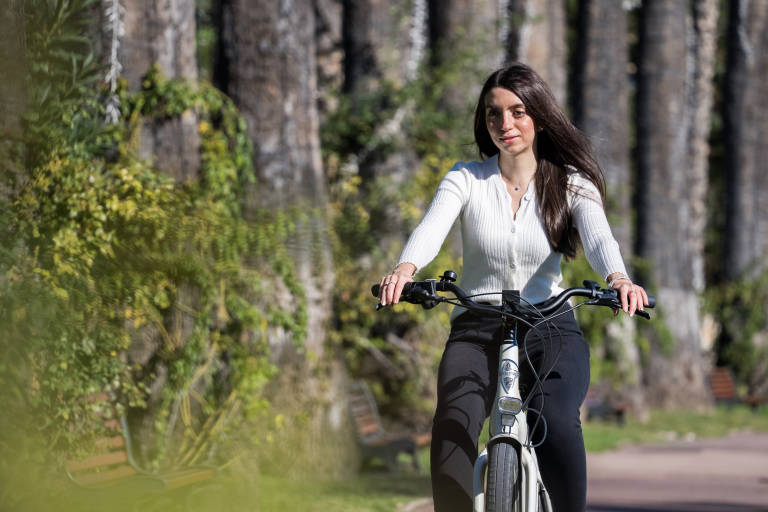 A woman in a white cardigan rides an electric bike through a park lined with tall trees and lush greenery.