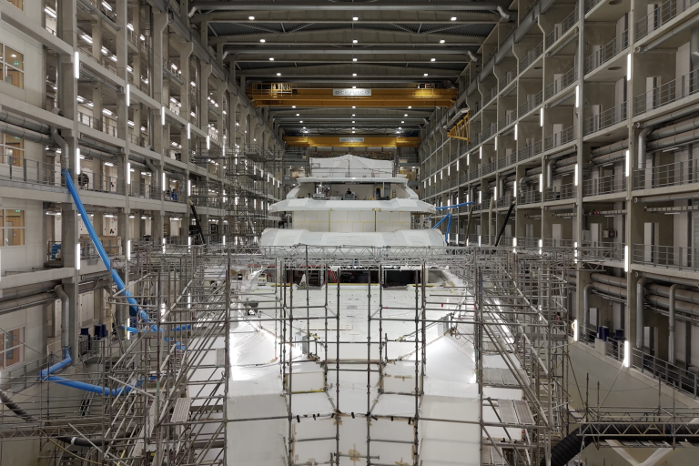 A massive yacht under construction is dwarfed by the immense scale of a shipyard's interior.