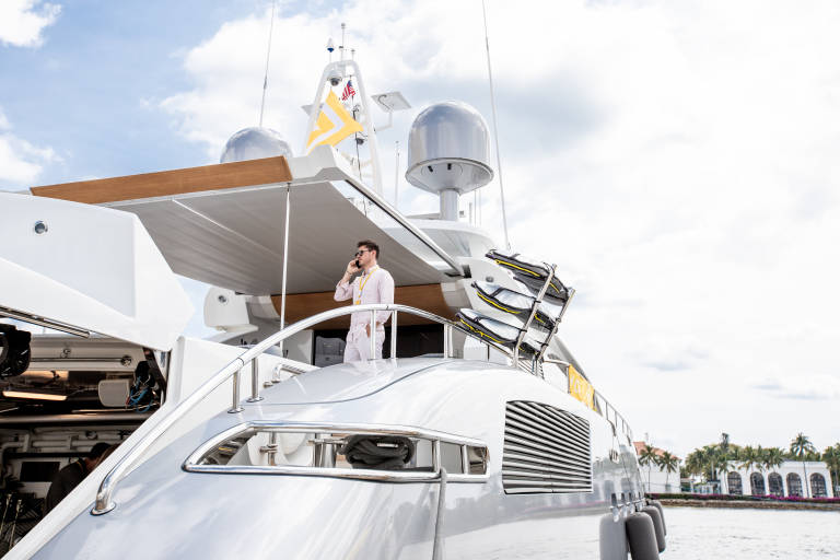 A man in a pink shirt talks on his phone while standing on the deck of a luxurious yacht.