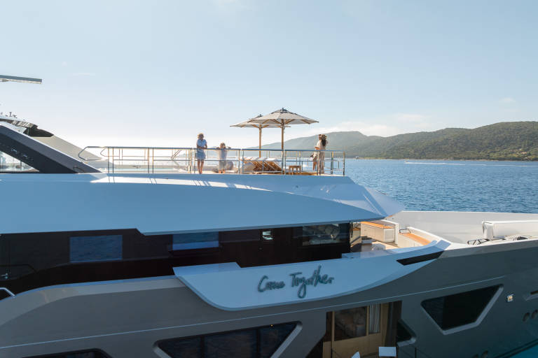 A luxurious yacht named "Come Together" glides through calm blue waters under a clear sky, with people enjoying the deck.