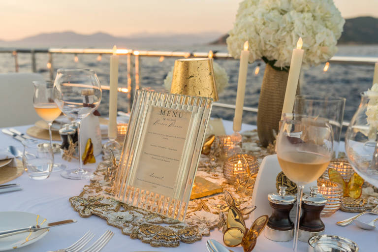 A beautifully set table awaits a Riviera dinner with a menu and candles as the sun sets over the water.