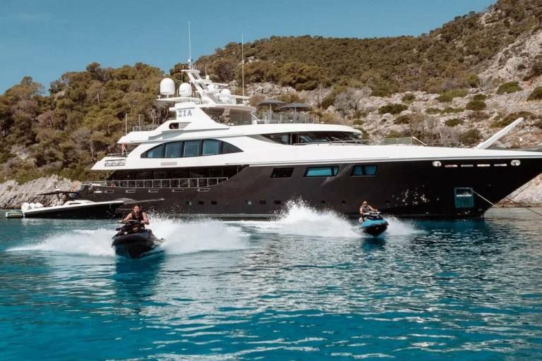 Two jet skiers race across the turquoise water near a luxurious yacht anchored by a rocky, tree-covered island.