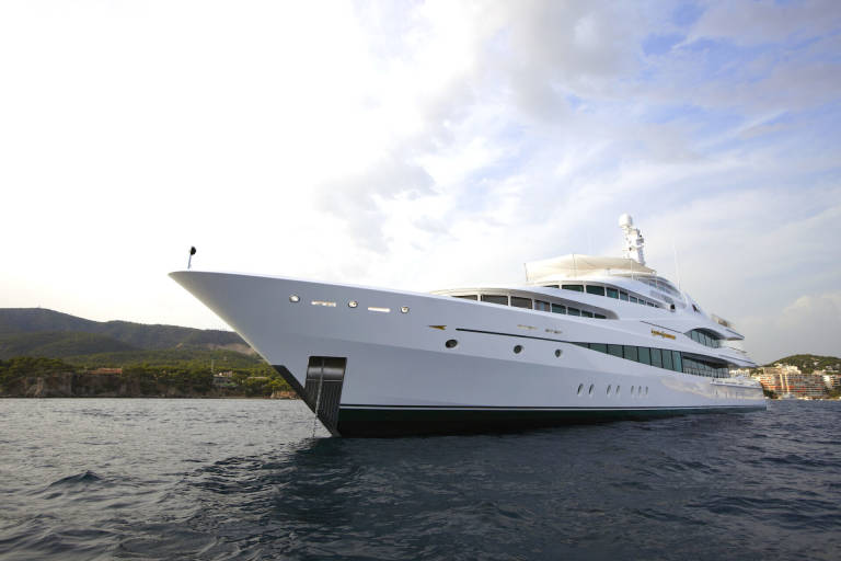 A luxurious white yacht glides through calm waters with a lush green coastline in the background.