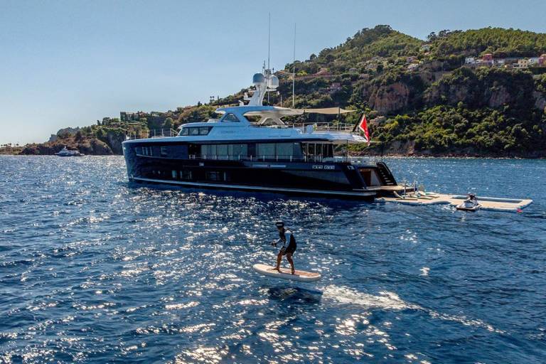 A person on a jet-powered surfboard glides across the sparkling blue water near a luxurious yacht anchored off a lush, green hillside.
