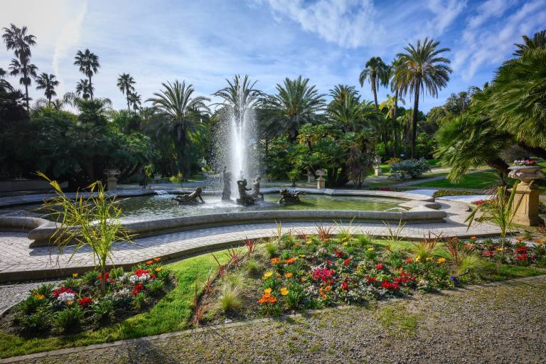 A vibrant fountain splashes in a lush garden surrounded by palm trees and colorful flowers under a bright blue sky.