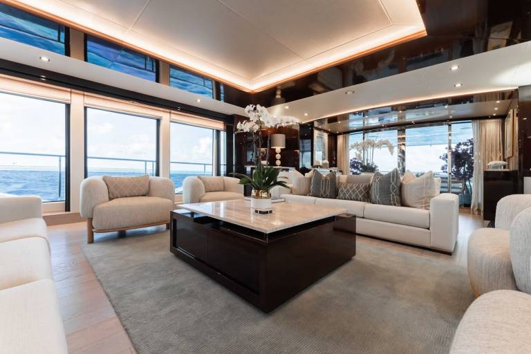 A luxurious and spacious living room with large windows offering a panoramic view of the ocean, featuring plush beige sofas, a dark wooden coffee table, and a modern lighting setup.