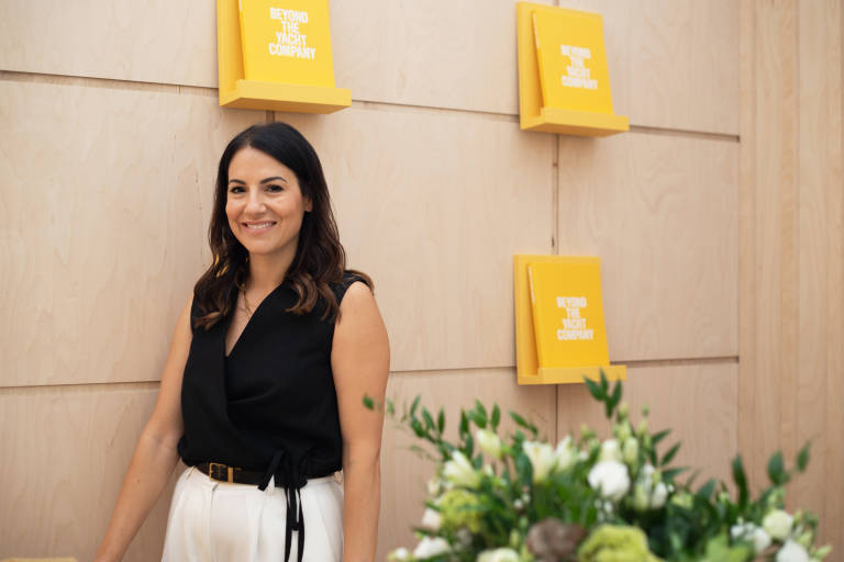 A smiling woman in a black top and white pants stands in front of a wood-paneled wall adorned with yellow signs.