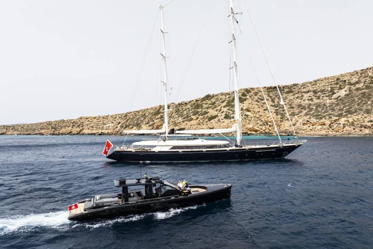 A sleek black tender navigates the choppy blue waters near a majestic sailing yacht anchored off a rocky, sun-drenched island.