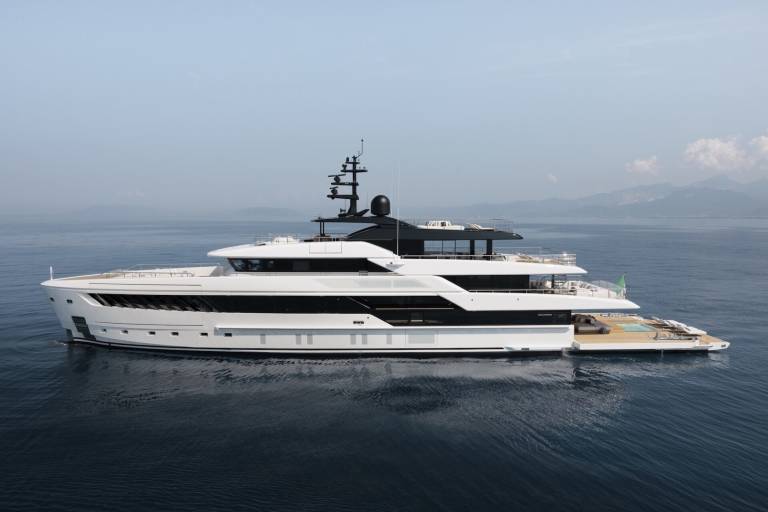 A sleek white superyacht glides through calm blue waters with a backdrop of hazy mountains.