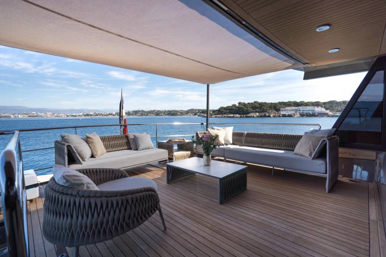 A modern yacht deck with comfortable seating in the foreground overlooks a calm blue sea and a distant coastline under a clear sky.