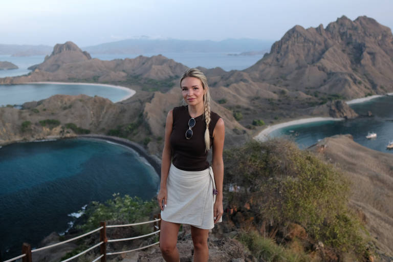 A blonde woman stands on a grassy hilltop in the foreground, with a stunning vista of rugged islands and turquoise bays in the background.