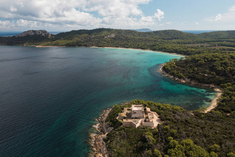 A stone fortress sits on a rocky point in the foreground, overlooking a turquoise bay surrounded by lush green hills and a distant mountain range under a cloudy sky.
