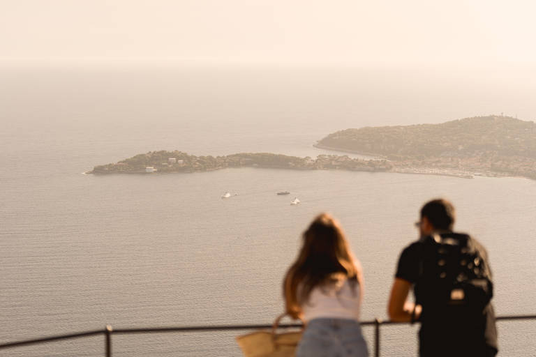 A couple stands at a railing in the foreground, gazing out at a hazy, sun-drenched bay dotted with sailboats and a distant, tree-covered peninsula.
