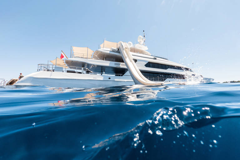 A large white yacht with a slide is anchored in clear blue water, with a rocky coastline visible in the distance under a bright sky.