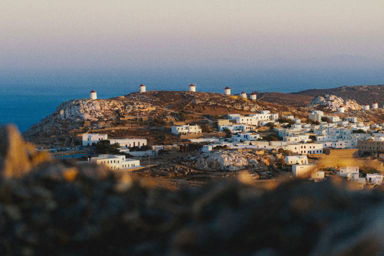 Rocky foreground frames a whitewashed village nestled on a hillside dotted with windmills, with the sea and a soft sky in the background.