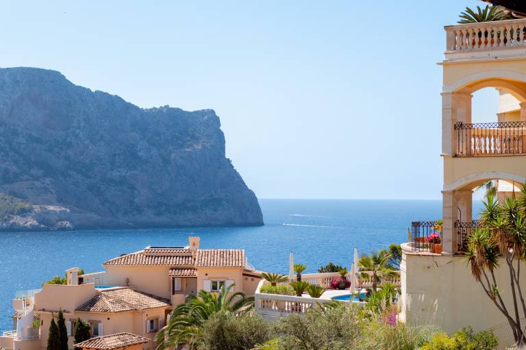 Terracotta-roofed villas with lush greenery in the foreground overlook a calm blue sea and a dramatic cliff face in the background.