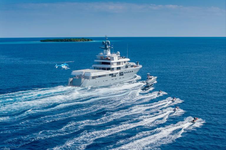 A large gray yacht cruises through the ocean, trailed by jet skis, with a helicopter and a small island in the distance under a clear blue sky.