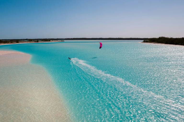 A kiteboarder glides across turquoise water, leaving a white wake, with a sandy shore on the left and a tree-lined horizon under a clear blue sky in the background.