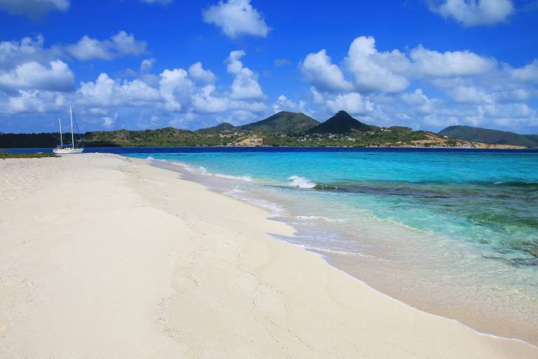 A pristine white sand beach curves towards turquoise waters, with a sailboat anchored in the distance against a backdrop of lush, green hills under a bright blue sky dotted with fluffy clouds.