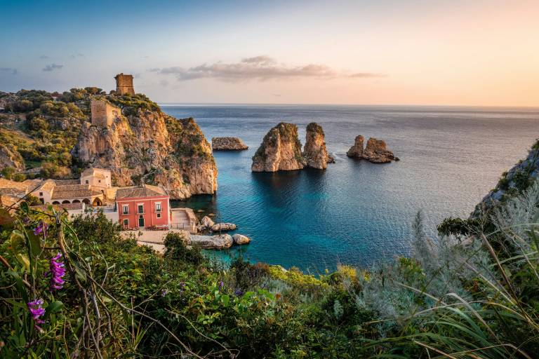 A pink building and ancient towers sit on a rocky coastline in the foreground, while rocky islets rise from the calm, blue sea under a soft, pastel sky in the background.
