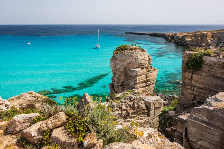 Rugged, sun-bleached rocks and sparse vegetation frame a vibrant turquoise sea with a sailboat in the distance.