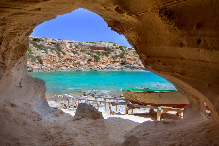 A weathered boat rests on wooden supports on a sandy shore within a cave, overlooking a vibrant turquoise sea and rocky cliffs under a clear blue sky.
