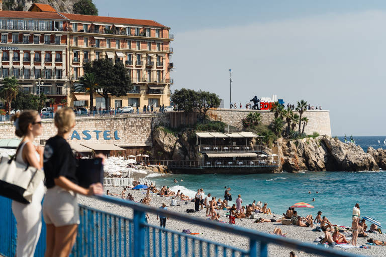 Two women stand on a blue railing overlooking a crowded pebble beach with a large hotel and rocky coastline in the background.