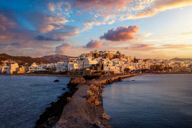 A stone pathway leads to a whitewashed village nestled on a hill overlooking the sea, under a colorful sunset sky.