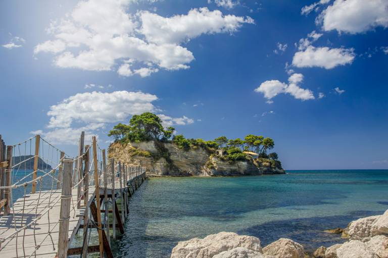 A wooden footbridge leads to a rocky island topped with trees, set against a clear blue sky with fluffy white clouds.