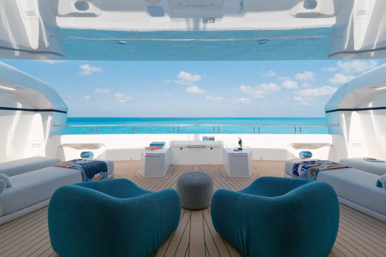 Two plush teal armchairs face a serene ocean view from a modern yacht deck, with white lounge chairs and a gray ottoman in the foreground and a bright blue sky with scattered clouds in the background.