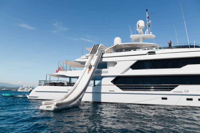A person slides down an inflatable water slide attached to a large white yacht, with other boats and a clear blue sky in the background.